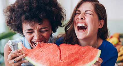 Two women, laughing, trying to eat a slice of watermelon